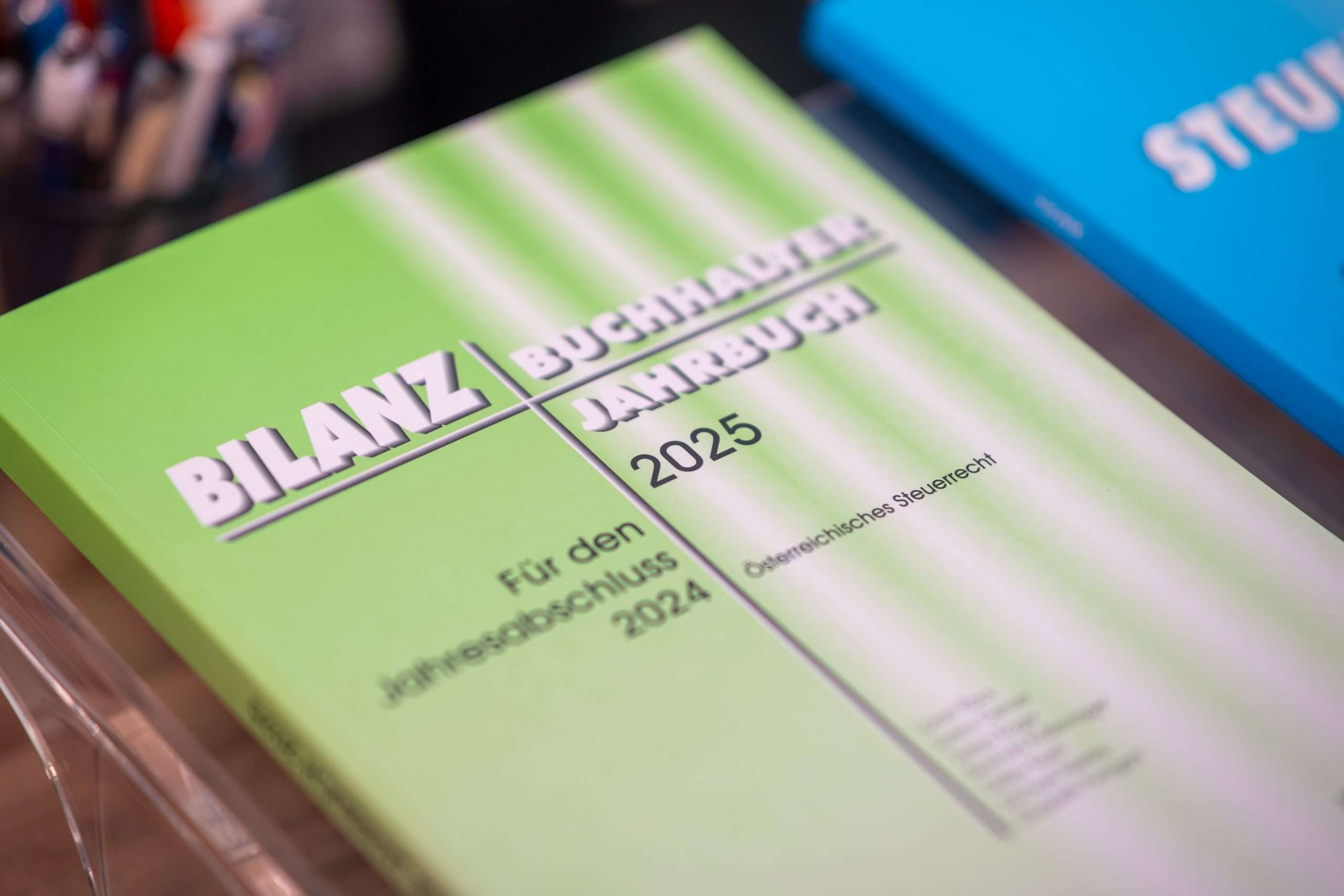 Nahaufnahme eines Grünbuchs mit dem Titel „BILANZ BUCHHALTER JAHRBUCH 2025“ von Birgit Hammer und dem Untertitel „Für den Jahresabschluss 2024“ mit den Schwerpunkten Rechnungslegung und Steuerrecht. Im Hintergrund ist teilweise ein weiteres blaues Buch zu sehen, möglicherweise von Bilanzbuchhalterin Gänserndorf.