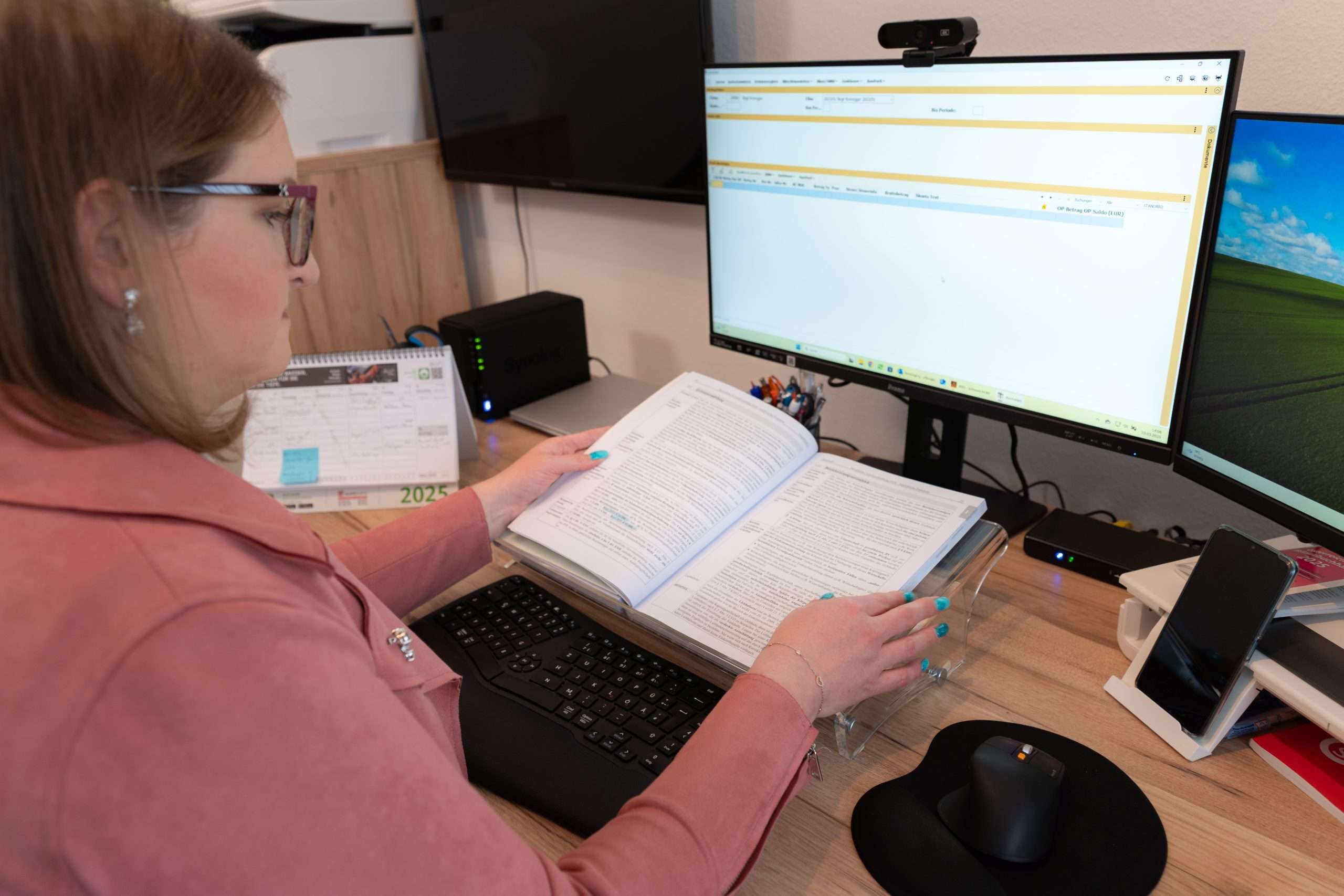 Eine Person mit Brille liest ein großes offenes Buch an einem Schreibtisch. Auf einem Computerbildschirm wird eine Webseite angezeigt. Der Arbeitsplatz, der für eine fleißige Bilanzbuchhalterin geeignet ist, verfügt über eine Tastatur, einen Kalender und einen Telefonständer. Die Person sitzt in einer rosa Jacke.
