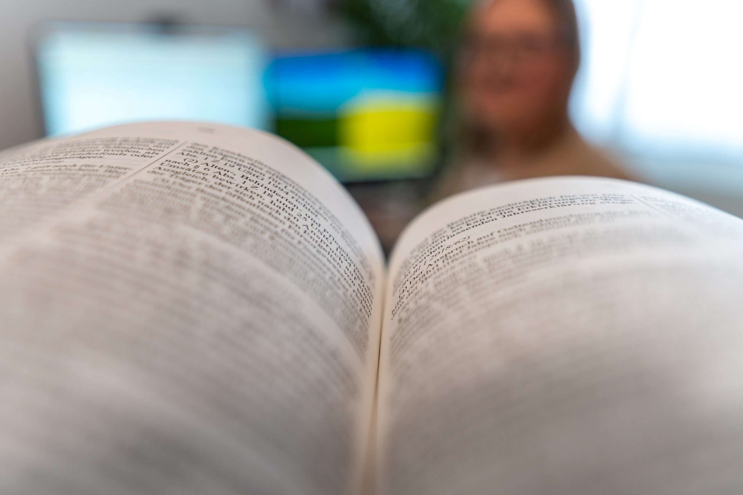 Im Vordergrund steht ein aufgeschlagenes Wörterbuch, dessen Text unscharf ist. Im unscharfen Hintergrund zeigt ein Computerbildschirm eine malerische Landschaft, während eine Bilanzbuchhalterin aus Gänserndorf an ihrem Schreibtisch Finanzdaten prüft und sich mit Präzision und Sachverstand auf den Jahresabschluss vorbereitet.