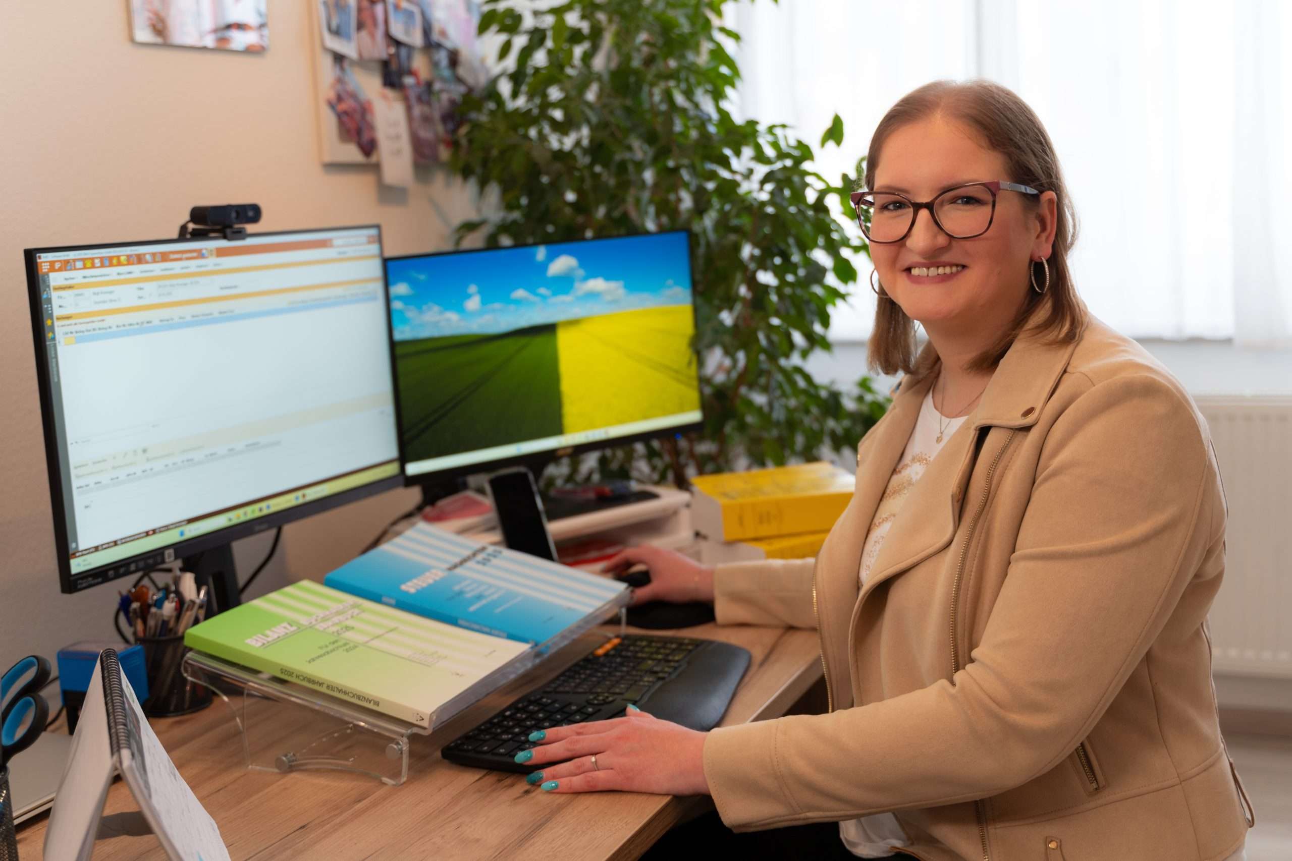 Birgit Hammer, engagierte Bilanzbuchhalterin, sitzt an ihrem Schreibtisch mit zwei Monitoren. Lächelnd erledigt sie Buchhaltungsaufgaben am Computer. Verschiedene Büroutensilien sind ordentlich um sie herum angeordnet, und eine große Pflanze gedeiht im natürlichen Licht, das den Raum durchflutet.