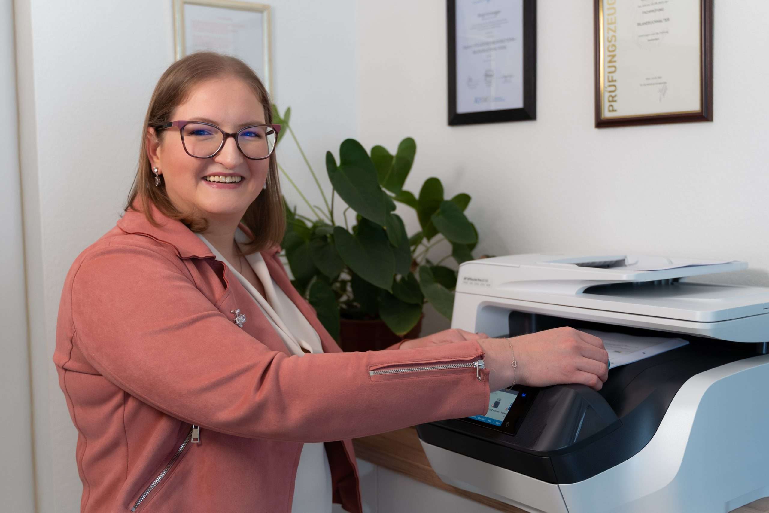 Eine Frau mit Brille und rosa Jacke steht lächelnd da und bedient einen Drucker. An der Wand hinter ihr hängen Zertifikate, die ihre Expertise als Bilanzbuchhalterin in Gänserndorf widerspiegeln. Eine Topfpflanze sorgt für einen Hauch von Natur neben dem Summen effizienter Buchhaltungstätigkeit.