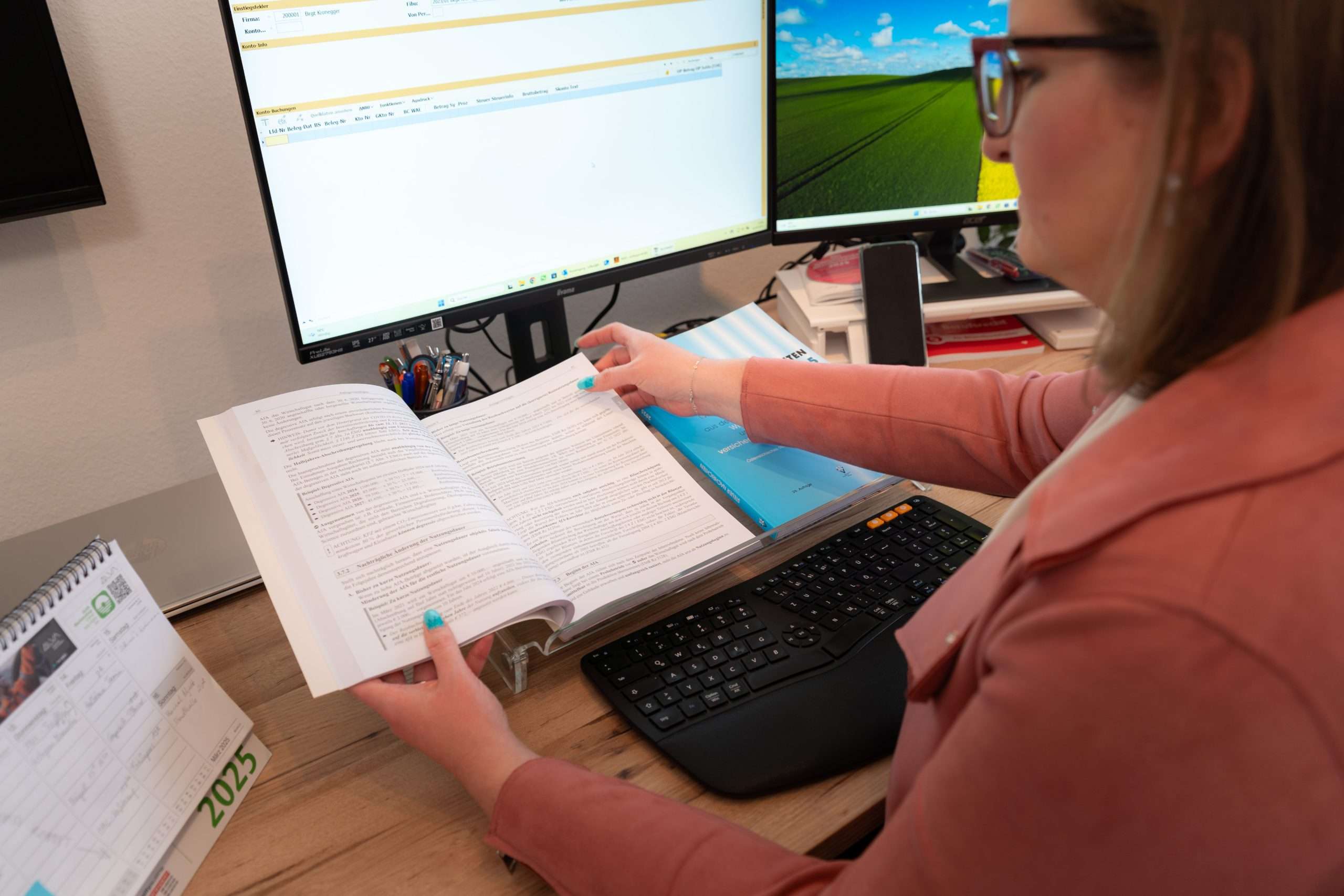 Eine Bilanzbuchhalterin mit Brille liest ein Buch über Buchhaltung und sitzt an einem Schreibtisch mit zwei Monitoren. Sie trägt eine rosa Jacke und blauen Nagellack. Der Schreibtisch ist mit Kalender, Tastatur und Schreibwaren ausgestattet – perfekt für die Vorbereitung des Jahresabschlusses.