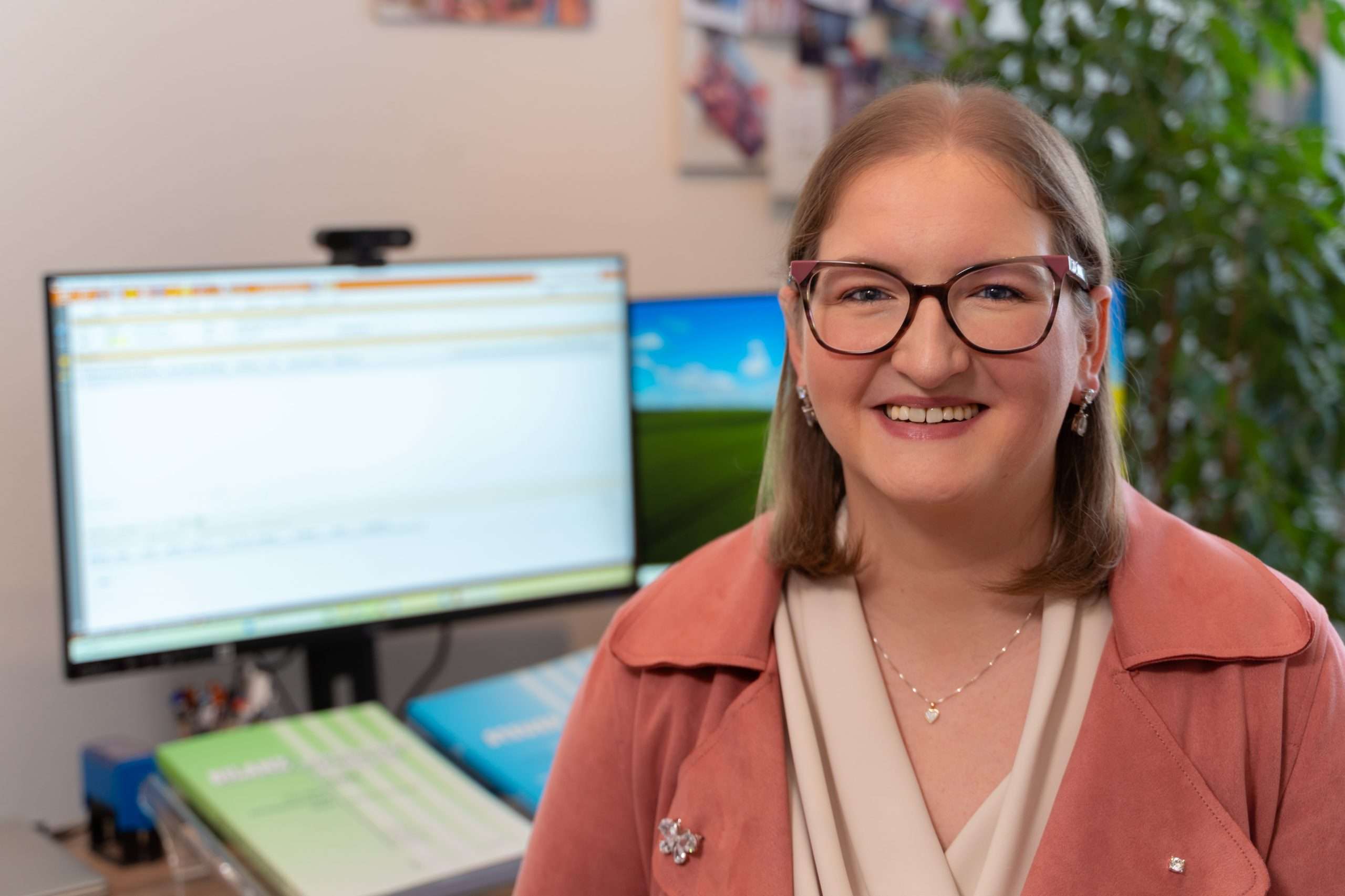 Eine Person mit schulterlangem Haar und Brille lächelt in die Kamera und verkörpert den professionellen und zugleich einladenden Charakter von Birgit Hammer. Sie trägt eine rosa Jacke über einem beigen Oberteil. Im Hintergrund ist ein Büroarbeitsplatz mit Computerbildschirm und Pflanzen zu sehen – perfekt für Beratung & Analyse.